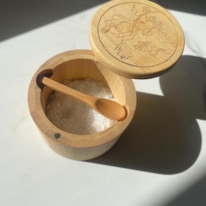 Op de afbeelding: Een bamboe zoutvaatje met een houten lepel. De ronde container heeft een deksel met een bloemenontwerp. De container is gevuld met wit zout. De lepel rust in de container.