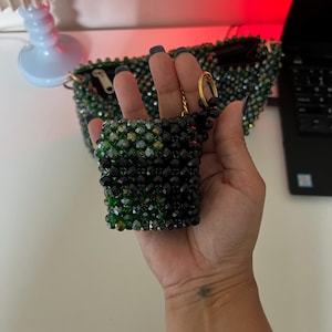 May include: A small, beaded purse in shades of green and black is held up. The purse is square-shaped and has a gold-colored ring attached to it. A larger beaded bag is in the background. The beads are small and reflective.