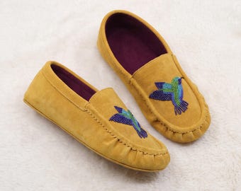 May include: A pair of mustard yellow suede moccasins featuring a beaded hummingbird design on the top of each shoe. The hummingbirds are crafted with blue, green, and purple beads. The interior of the shoes is lined with a deep purple fabric.