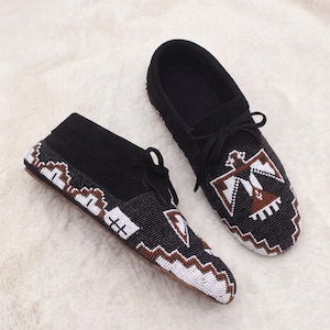 May include: A pair of black suede moccasins featuring intricate beadwork. The shoes are decorated with white, black, and brown geometric patterns and a central bird motif on the vamp, complete with black laces and a flat sole.