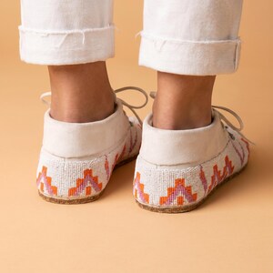 Pu&ograve; includere: Un paio di mocassini bianchi con intricate perline in motivi geometrici arancioni, rosa e viola. Le scarpe presentano una silhouette bassa con lacci bianchi e una suola piatta di colore chiaro, viste dal retro su uno sfondo neutro.