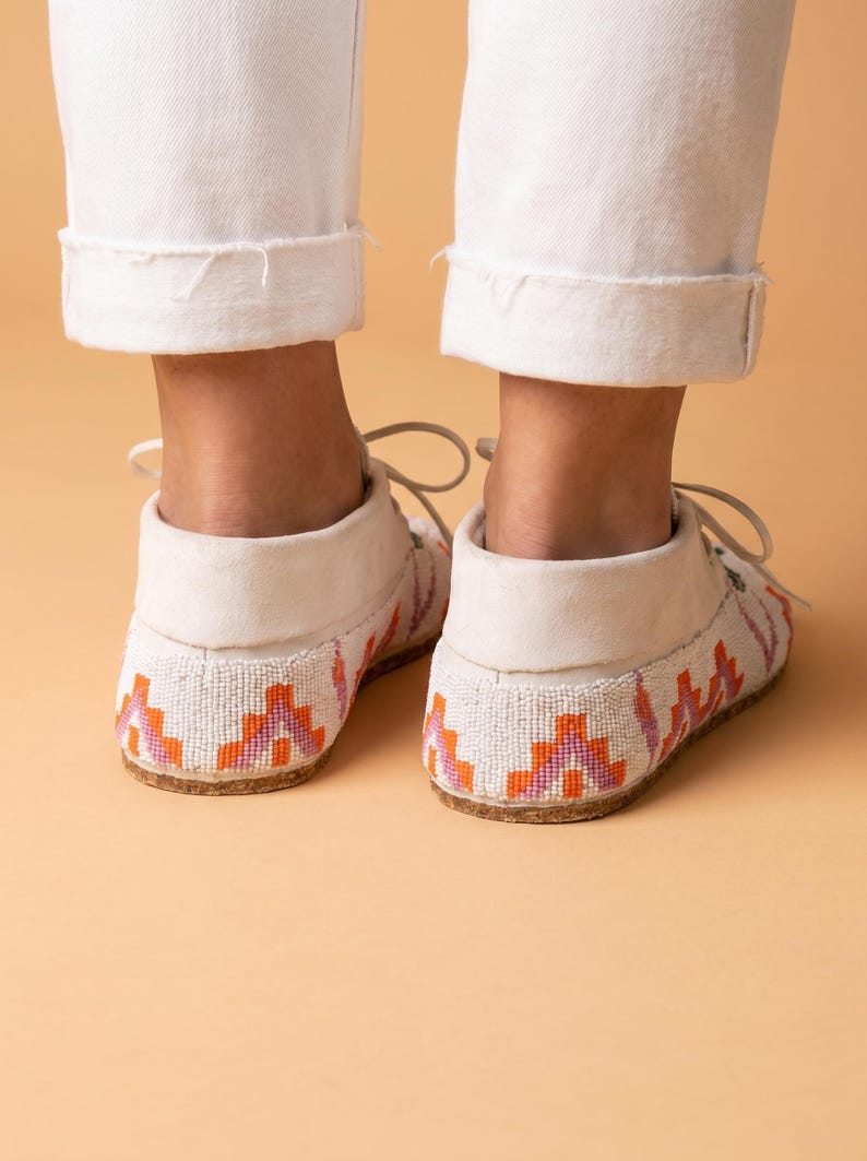 Pu&ograve; includere: Un paio di mocassini bianchi con intricate perline in motivi geometrici arancioni, rosa e viola. Le scarpe presentano una silhouette bassa con lacci bianchi e una suola piatta di colore chiaro, viste dal retro su uno sfondo neutro.
