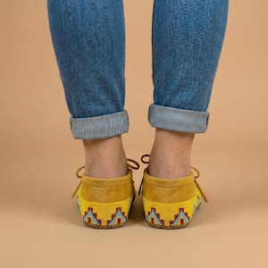 Pu&ograve; includere: Un paio di mocassini in camoscio giallo con un motivo geometrico di perline rosso, azzurro e blu scuro sul tallone. Le scarpe hanno sottili lacci gialli annodati a fiocco e sono indossate con jeans in denim blu con risvolto.