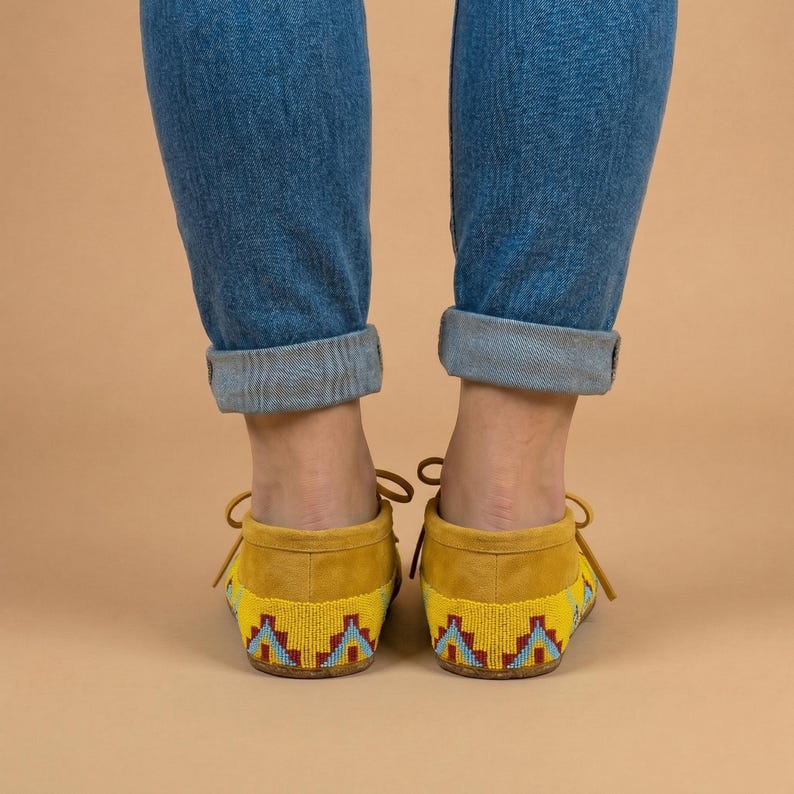 Pu&ograve; includere: Un paio di mocassini in camoscio giallo con un motivo geometrico di perline rosso, azzurro e blu scuro sul tallone. Le scarpe hanno sottili lacci gialli annodati a fiocco e sono indossate con jeans in denim blu con risvolto.