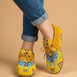 Pu&ograve; includere: Mocassini gialli con intricate perline in motivi geometrici blu, rossi e neri. Queste scarpe stringate presentano una consistenza morbida simile alla pelle scamosciata sul tallone e sul colletto, con punta arrotondata e sottili lacci in pelle gialla.