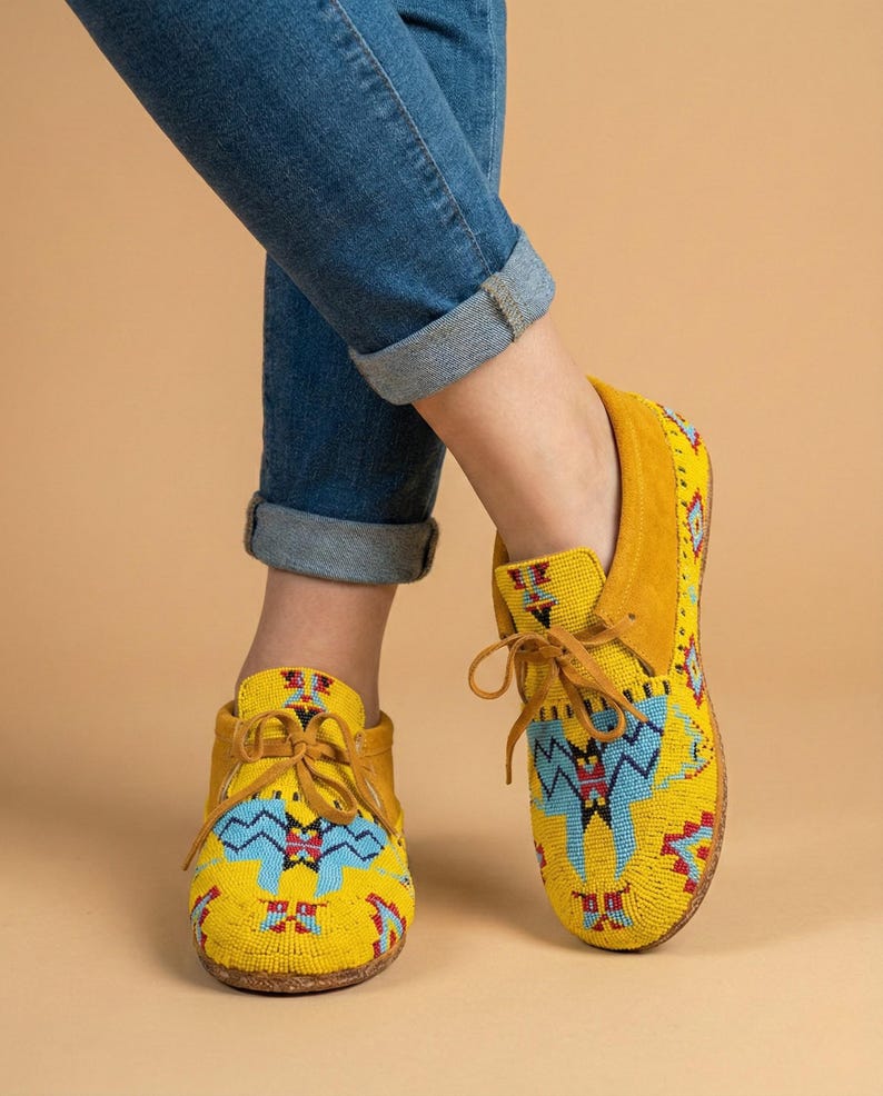 Pu&ograve; includere: Mocassini gialli con intricate perline in motivi geometrici blu, rossi e neri. Queste scarpe stringate presentano una consistenza morbida simile alla pelle scamosciata sul tallone e sul colletto, con punta arrotondata e sottili lacci in pelle gialla.