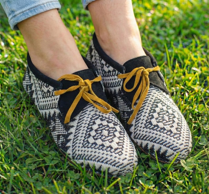 May include: Pair of black and white patterned shoes with yellow laces. The shoes have a geometric design and a black suede collar. The shoes are on a bed of green grass.