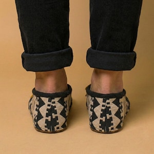 May include: A pair of patterned slippers with a black and tan geometric design. The slippers are worn with black corduroy pants. The background is a solid tan color. The slippers appear to be made of a soft material.