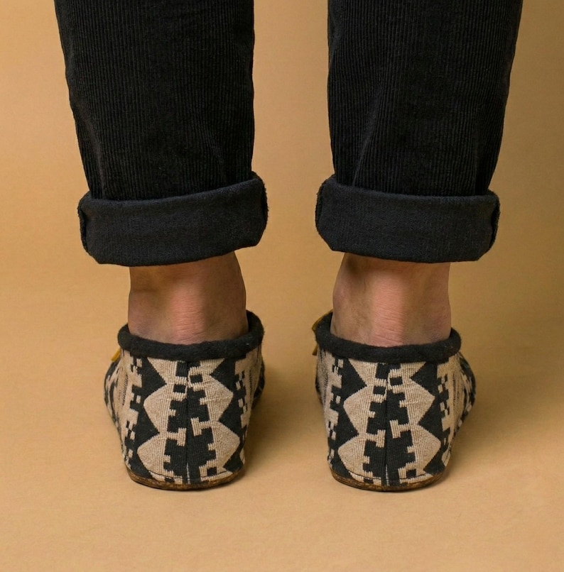 May include: A pair of patterned slippers with a black and tan geometric design. The slippers are worn with black corduroy pants. The background is a solid tan color. The slippers appear to be made of a soft material.