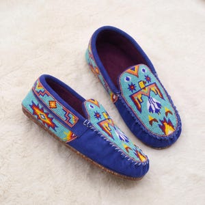 May include: A pair of blue moccasins featuring intricate beadwork in geometric and avian patterns. The shoes are crafted with a vibrant light blue beaded vamp and side panels, contrasted by a deep blue base and a brown textured sole.
