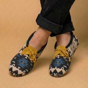 May include: Close-up of a pair of patterned moccasin-style shoes. The shoes feature a black, blue, and beige geometric design with a yellow suede tongue and laces. The shoes are worn with black corduroy pants.