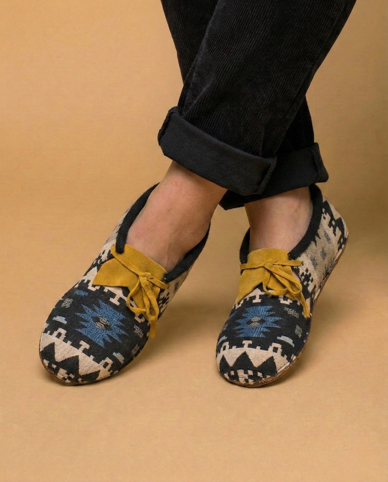 May include: Close-up of a pair of patterned moccasin-style shoes. The shoes feature a black, blue, and beige geometric design with a yellow suede tongue and laces. The shoes are worn with black corduroy pants.