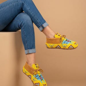 Pu&ograve; includere: Un paio di mocassini gialli caratterizzati da un vivace motivo geometrico di perline nei toni del blu, rosso e bianco. Le scarpe presentano inserti in camoscio color cuoio sul tallone e sul colletto, con lacci abbinati annodati sul davanti.