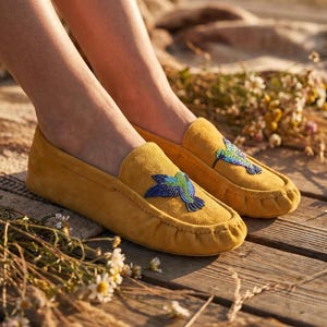 May include: A pair of mustard yellow suede moccasins featuring a beaded hummingbird design on the top of each shoe. The hummingbird is crafted with blue, green, and purple beads, and the shoes have a traditional stitched toe seam construction.