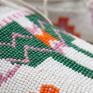Pu&ograve; includere: Un primo piano di un mocassino in pelle bianca caratterizzato da intricate perline con un motivo geometrico. Il design incorpora piccole perline sferiche in tonalit&agrave; vivaci di verde bosco, arancione brillante e rosa tenue su fondo bianco.