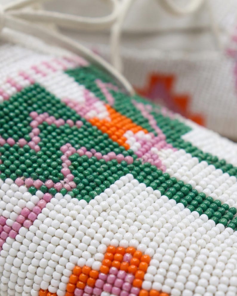 Pu&ograve; includere: Un primo piano di un mocassino in pelle bianca caratterizzato da intricate perline con un motivo geometrico. Il design incorpora piccole perline sferiche in tonalit&agrave; vivaci di verde bosco, arancione brillante e rosa tenue su fondo bianco.