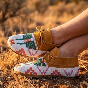 Könnte beinhalten: Ein Paar hellbraune Wildleder-Mokassins mit aufwendiger Perlenarbeit. Die weißen, mit Perlen besetzten Oberteile weisen geometrische Muster in Grün, Orange und Rosa auf. Die Schuhe haben hellbraune Lederschnürsenkel.