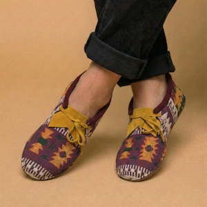 May include: A pair of patterned slip-on shoes with a burgundy and yellow design. The shoes have a yellow suede tongue and laces. The shoes are worn with black corduroy pants.