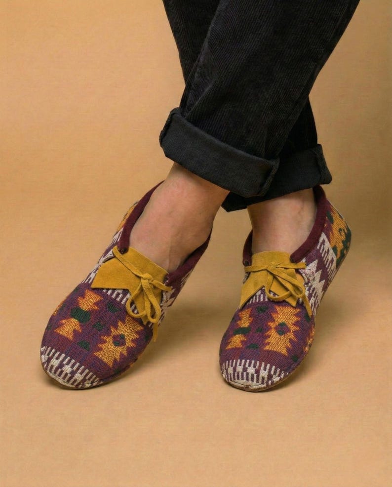 May include: A pair of patterned slip-on shoes with a burgundy and yellow design. The shoes have a yellow suede tongue and laces. The shoes are worn with black corduroy pants.