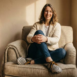 May include: A woman seated in a beige armchair, wearing a cream-colored cable-knit cardigan, blue jeans, and patterned moccasin-style slippers. A beige blanket is draped over the arm of the chair. The background is a neutral wall with natural light.