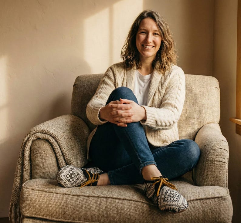 May include: A woman seated in a beige armchair, wearing a cream-colored cable-knit cardigan, blue jeans, and patterned moccasin-style slippers. A beige blanket is draped over the arm of the chair. The background is a neutral wall with natural light.