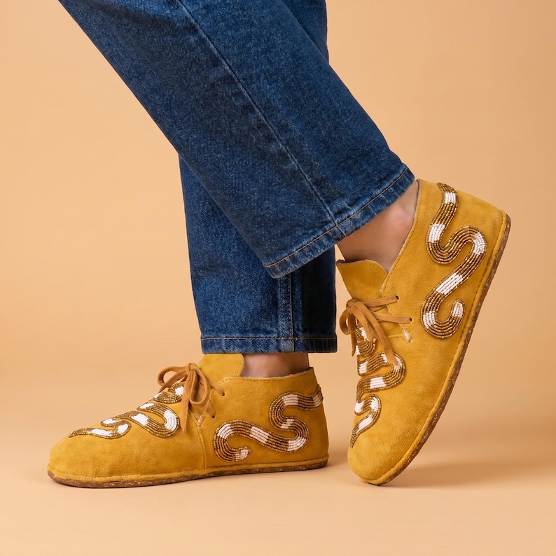 May include: A pair of mustard yellow suede lace-up shoes featuring a decorative snake pattern created with small brown and white beads. The shoes have a low-top silhouette with matching yellow laces and a flat, textured sole for a casual appearance.