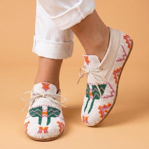 Pu&ograve; includere: Un paio di mocassini bianchi caratterizzati da intricate perline con motivi geometrici verdi, arancioni e viola. Le scarpe presentano una chiusura frontale con lacci sottili in pelle bianca e una suola chiara, con un dettagliato design a perline.