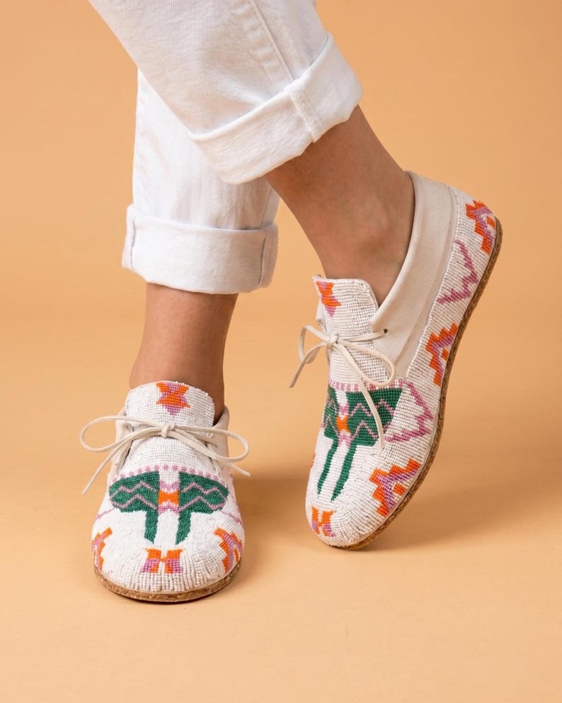 Pu&ograve; includere: Un paio di mocassini bianchi caratterizzati da intricate perline con motivi geometrici verdi, arancioni e viola. Le scarpe presentano una chiusura frontale con lacci sottili in pelle bianca e una suola chiara, con un dettagliato design a perline.