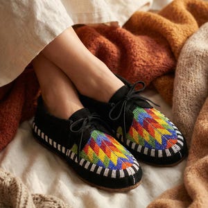 Op de afbeelding: Zwarte su&egrave;de mocassins met kleurrijke, geometrische kralenpatronen. De schoenen hebben zwarte veters en witte kralenranden. De afbeelding toont de schoenen op een bed met diverse dekens.