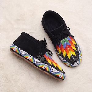 May include: A pair of black suede moccasins featuring intricate beadwork in a vibrant rainbow geometric pattern. The shoes include black laces and a tan sole, with the colorful beadwork extending along the sides and across the toe box.