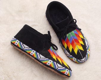 May include: A pair of black suede moccasins featuring intricate beadwork in a vibrant rainbow geometric pattern. The shoes include black laces and a tan sole, with the colorful beadwork extending along the sides and across the toe box.
