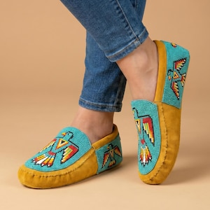 May include: Mustard yellow suede moccasins featuring turquoise beaded panels on the vamp and sides. The beadwork displays a stylized bird motif in vibrant red, orange, yellow, and black colors. The slip-on shoes have a soft, flexible construction.