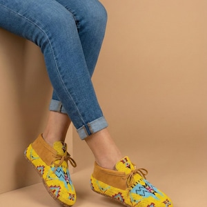 Pu&ograve; includere: Un paio di mocassini gialli con intricate perline in motivi geometrici blu, rossi e bianchi, abbinati a jeans blu. Le scarpe presentano inserti in camoscio beige sul tallone e sottili lacci in pelle marrone annodati sul davanti.