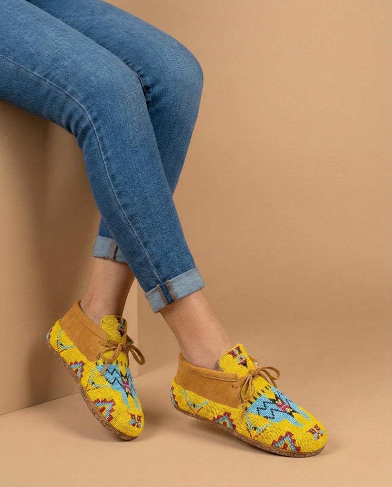 Pu&ograve; includere: Un paio di mocassini gialli con intricate perline in motivi geometrici blu, rossi e bianchi, abbinati a jeans blu. Le scarpe presentano inserti in camoscio beige sul tallone e sottili lacci in pelle marrone annodati sul davanti.