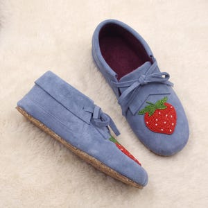 May include: A pair of light blue moccasins featuring a beaded strawberry design on the top. The shoes have a soft, suede-like texture, a dark purple interior lining, and a light-colored, textured sole with a tie closure at the front.