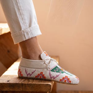 Pu&ograve; includere: Un mocassino bianco con intricate perline in motivi geometrici arancioni, rosa e verdi. La scarpa presenta un colletto in pelle bianca liscia, un sottile laccio sulla parte superiore e una suola piatta, indossata con pantaloni bianchi con risvolto.