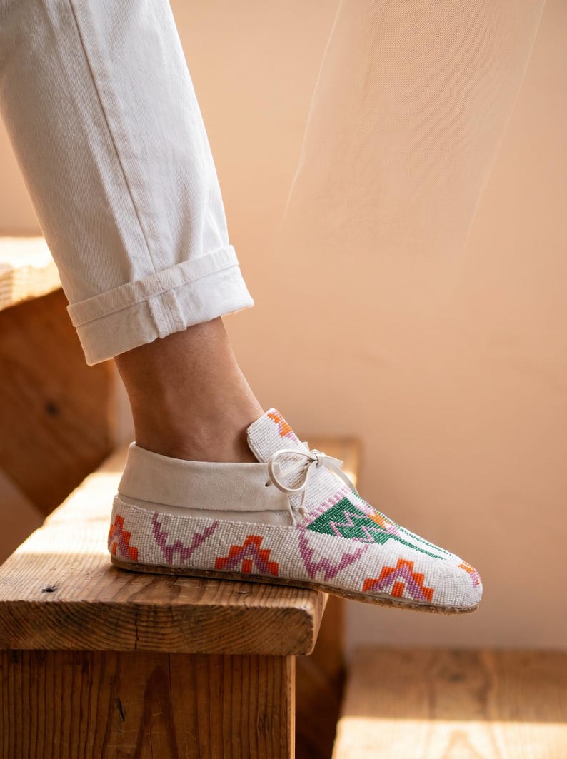 Pu&ograve; includere: Un mocassino bianco con intricate perline in motivi geometrici arancioni, rosa e verdi. La scarpa presenta un colletto in pelle bianca liscia, un sottile laccio sulla parte superiore e una suola piatta, indossata con pantaloni bianchi con risvolto.