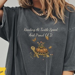 May include: Dark gray t-shirt with the text "Reading At Turtle Speed And Proud Of It." The shirt features a turtle with flowers on its back and butterflies. The shirt has a relaxed fit.