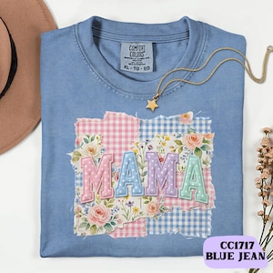 以下が含まれることがあります： パステルカラーの「MAMA」の文字が入った、ライトブルーのComfort Colors Tシャツ。文字は、花柄とギンガムチェックのパッチワークデザインになっています。星のペンダントが付いたゴールドのネックレスがシャツの上にあります。「CC1717 BLUE JEAN」の文字が下にあります。