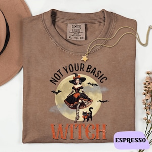 Puede incluir: Camiseta color espresso con un diseño de una bruja con vestido negro y sombrero, con el texto "NOT YOUR BASIC WITCH". Un collar dorado con forma de estrella está sobre la camiseta. La etiqueta dice "Espresso".