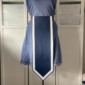 Puede incluir: Un vestido azul con un panel azul oscuro en la parte delantera, acentuado con rayas blancas. El vestido tiene un cinturón marrón y un dobladillo decorativo. El vestido se exhibe en un maniquí.