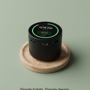 May include: A black cylindrical digital clock with a circular screen displaying 12:16 PM, the date, and prayer times. The device features a glowing green ring light, a side-mounted port, and a small indicator light, resting on a round wooden base.
