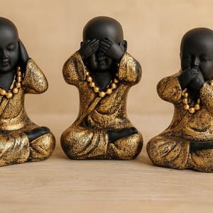 Op de afbeelding: Drie zwart-gouden Boeddha-beelden in de pose 'Zie geen kwaad, hoor geen kwaad, spreek geen kwaad'. Elk beeld heeft een zwart hoofd en handen, met een gouden gewaad en een gouden kralenketting. De beelden staan op een lichtbruin houten oppervlak.