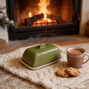 Peut inclure: Un beurrier en céramique vert avec un couvercle, posé sur une surface texturée de couleur crème. Une tasse de chocolat chaud et deux biscuits aux pépites de chocolat sont à proximité. Une cheminée avec un feu brûlant est en arrière-plan.