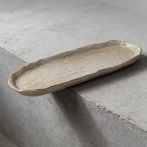 Può includere: Un vassoio da portata ovale in ceramica beige con una superficie strutturata e un bordo leggermente irregolare e ondulato. Il vassoio è posto su uno sfondo grigio neutro, che mette in risalto il suo design minimalista.