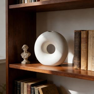 以下が含まれることがあります： 木製の棚に飾られた、白いセラミック製のドーナツ型花瓶。円形の開口部があります。小さな胸像の像といくつかの古い本も棚にあります。花瓶は滑らかでマットな仕上がりです。