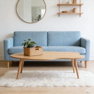 May include: A light blue sofa with a wooden coffee table in front of it. The table has a unique, organic shape and is supported by angled legs. A woven basket with a plant sits on the table. A white rug and a round mirror complete the scene.