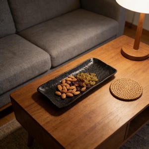 May include: A black ceramic serving dish with almonds, raisins, and golden raisins. A woven coaster sits to the right of the dish on a wooden coffee table. A grey sofa and a lamp are in the background.