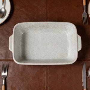 May include: A rectangular, light grey ceramic baking dish with speckled detailing and two side handles. The dish is surrounded by cutlery and a white plate on a brown leather surface.