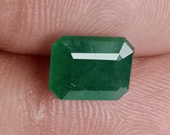 May include: A rectangular emerald gemstone with a deep green hue. The gem has a faceted cut with a rectangular shape and beveled edges. The emerald is held between two fingers, showcasing its clarity and color.